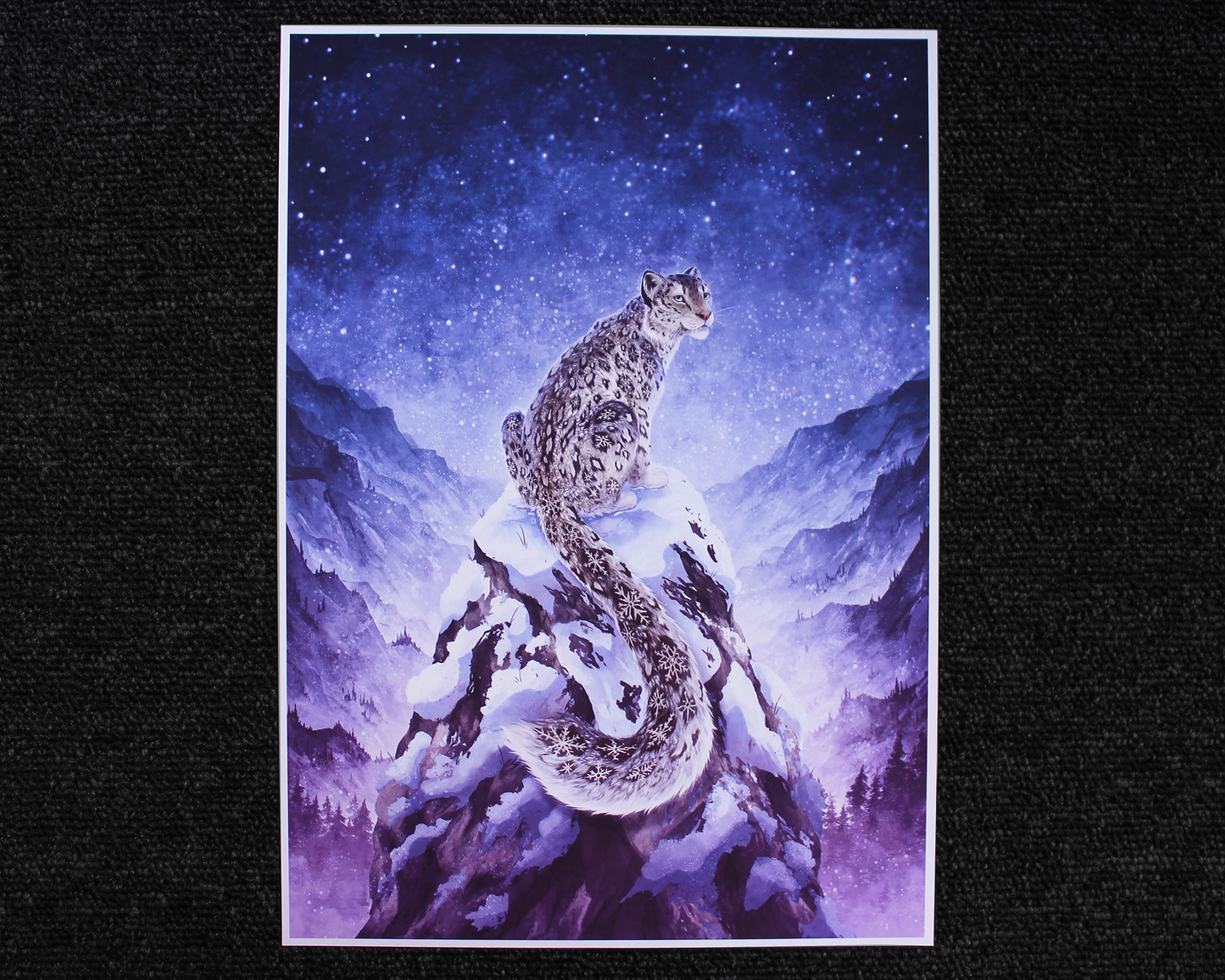 A4 Print of a snow leopard sat on the peak of a mountain. The snow leopard has snowflakes for the spot patterns, and in the background is a mountain range with a blue starry night sky.