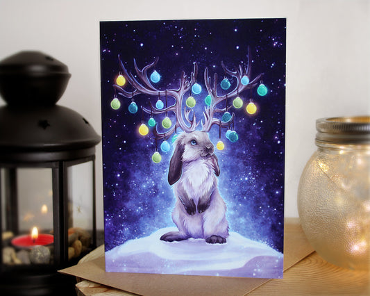 christmas greeting card of a festive jackalope, based off a lop-eared bunny rabbit. On their antlers hangs  baubles in various colours. The are sat on snow with a blue night starry sky backround.